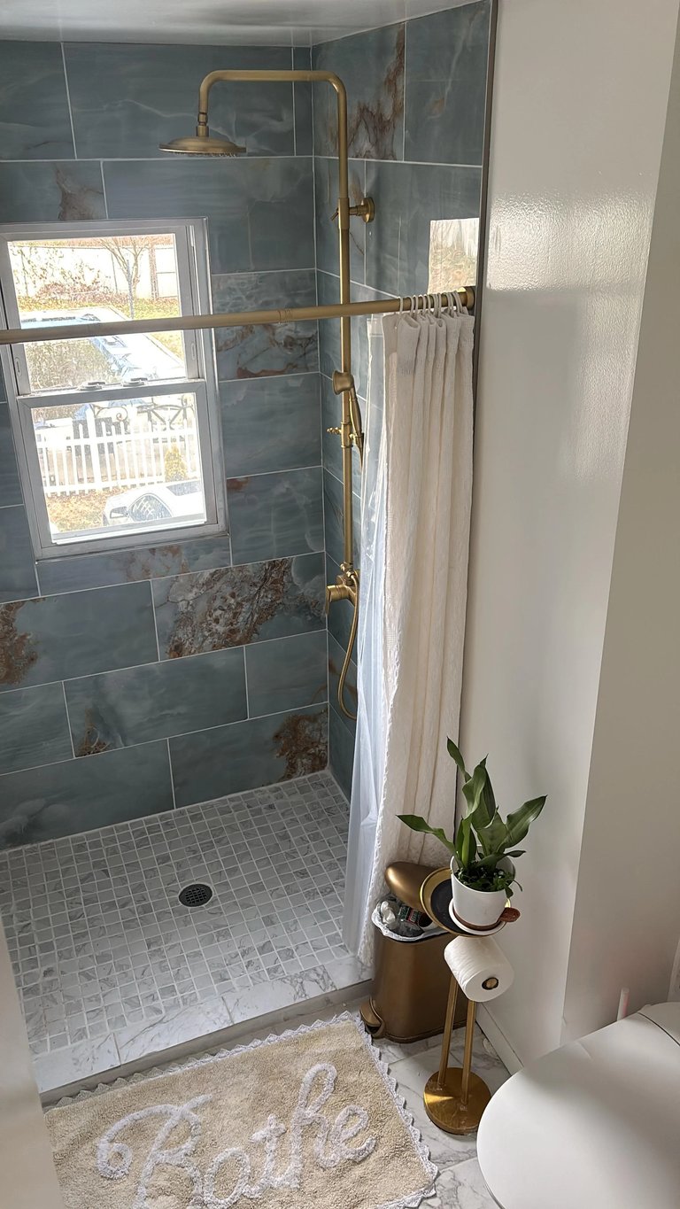 Modern bathroom shower with blue-gray tile walls, glass enclosure, brass fixtures, white curtain, and window view overlooking trees