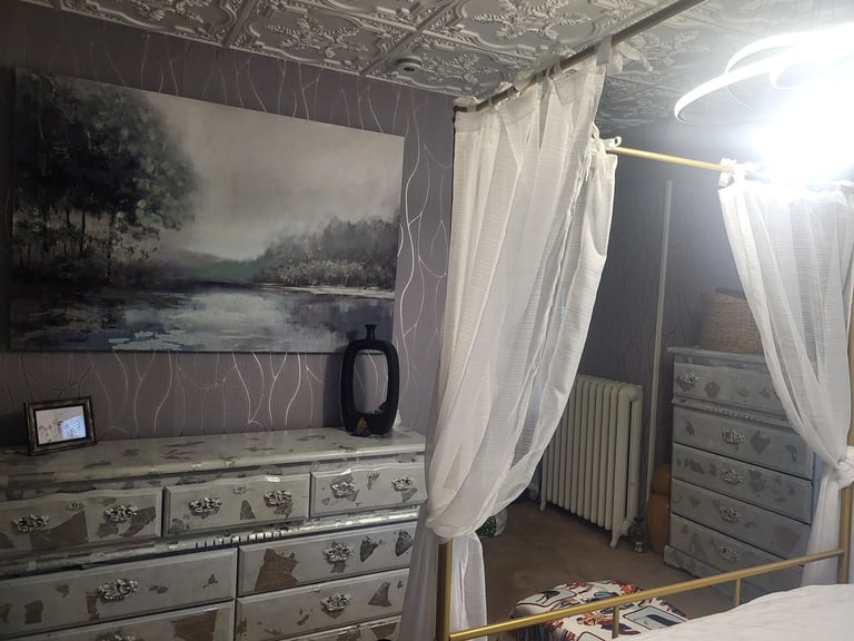 Cozy bedroom with sheer white curtains, ornate dresser, landscape artwork, and decorative lighting fixtures