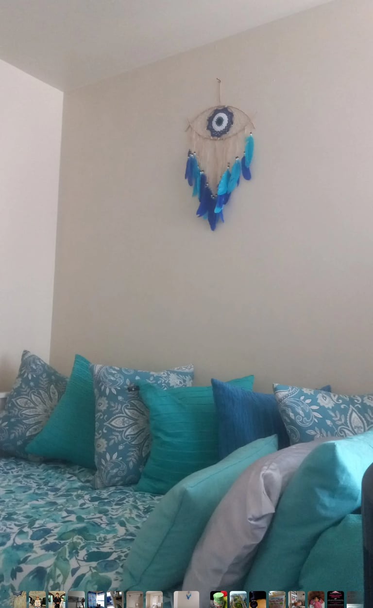 Blue feather dreamcatcher with evil eye design hanging on white bedroom wall above turquoise and patterned pillows