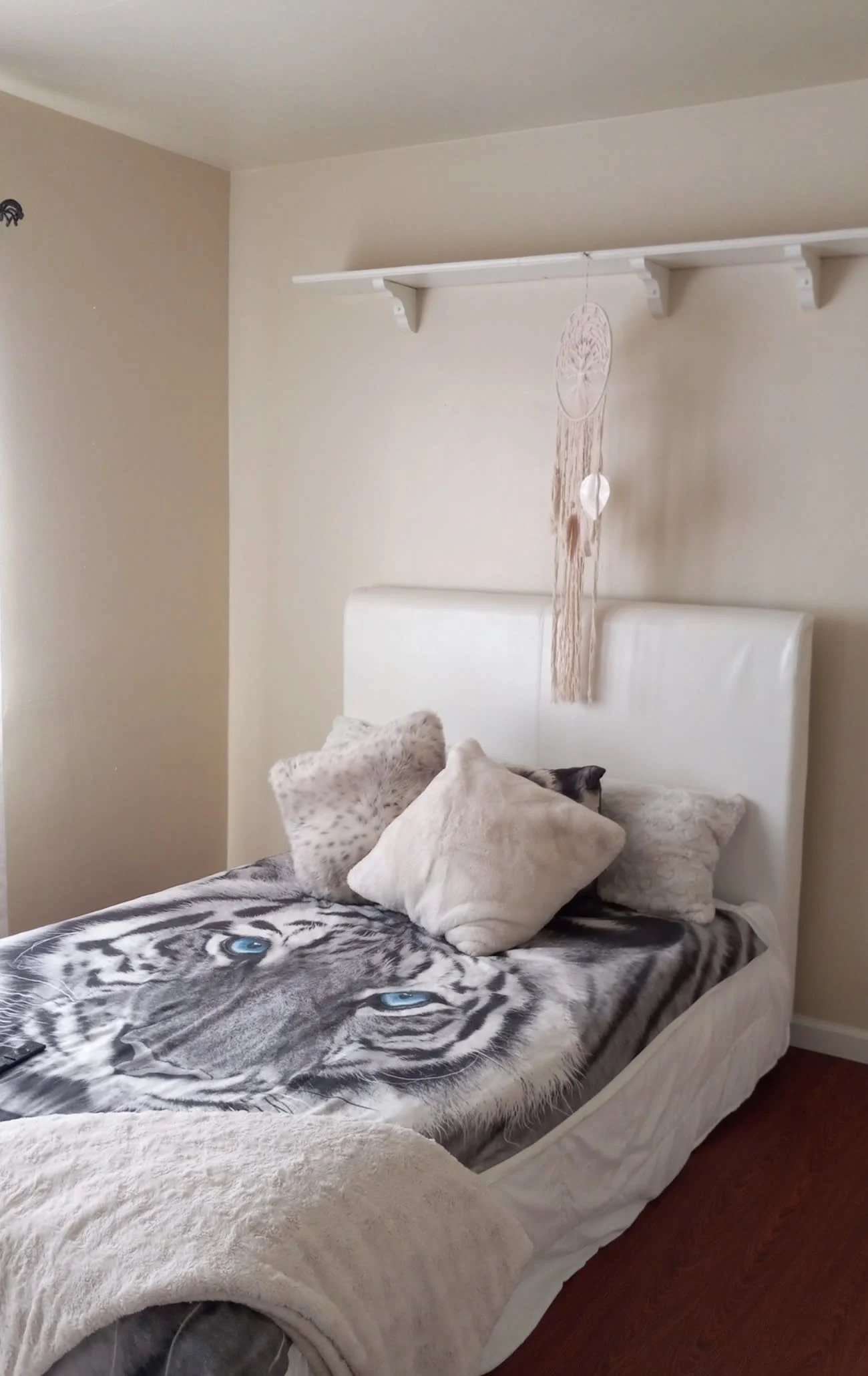 Minimalist bedroom with white upholstered bed, patterned bedding, decorative pillows, macrame wall hanging above headboard, and wooden flooring