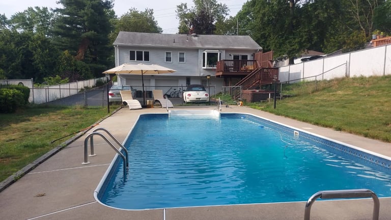Backyard in-ground swimming pool with two white deck chairs and house with trees in background